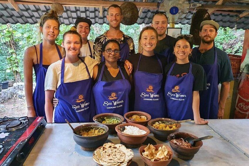 En gruppe mennesker i blå forklæder med teksten 'Sea Waves Cooking Class', der står rundt om et bord med forskellige fødevarer og køkkenredskaber. 1