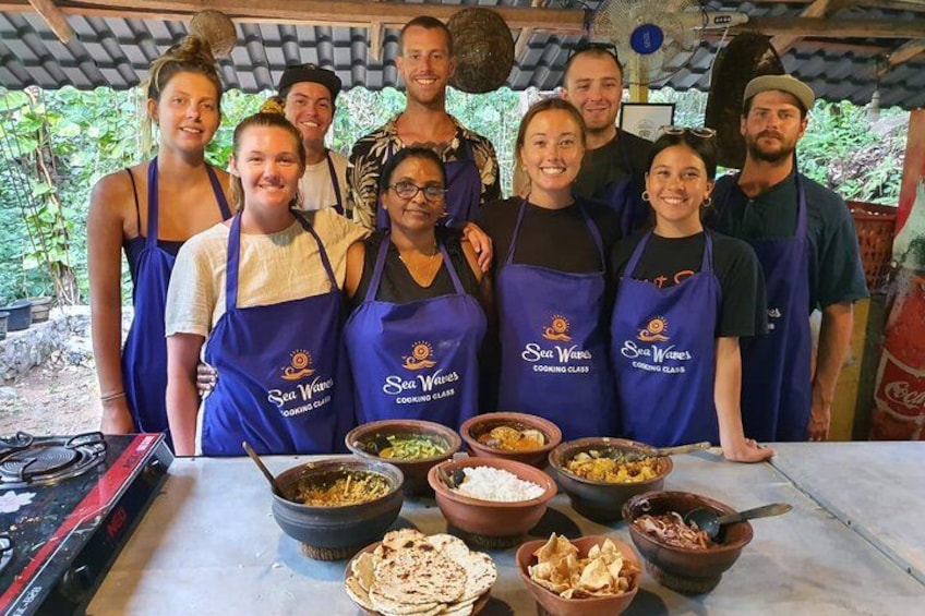 Un grupo de personas con delantales con el logotipo 'Sea Waves Cooking Class' reunidas alrededor de una mesa con diversos alimentos y utensilios de cocina. 1