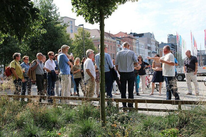 Un groupe de personnes se tient sur une zone pavée, avec de l’herbe et des plantes au premier plan, des bâtiments en arrière-plan et des drapeaux sur des mâts. 2