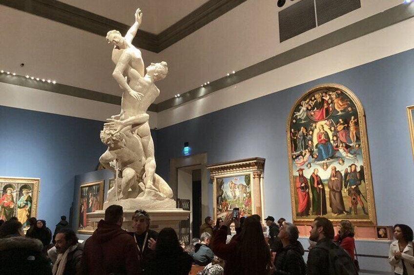 En marmorskulptur af en mandlig figur i en dynamisk positur, omgivet af besøgende i et museum med klassiske malerier på væggene. 4