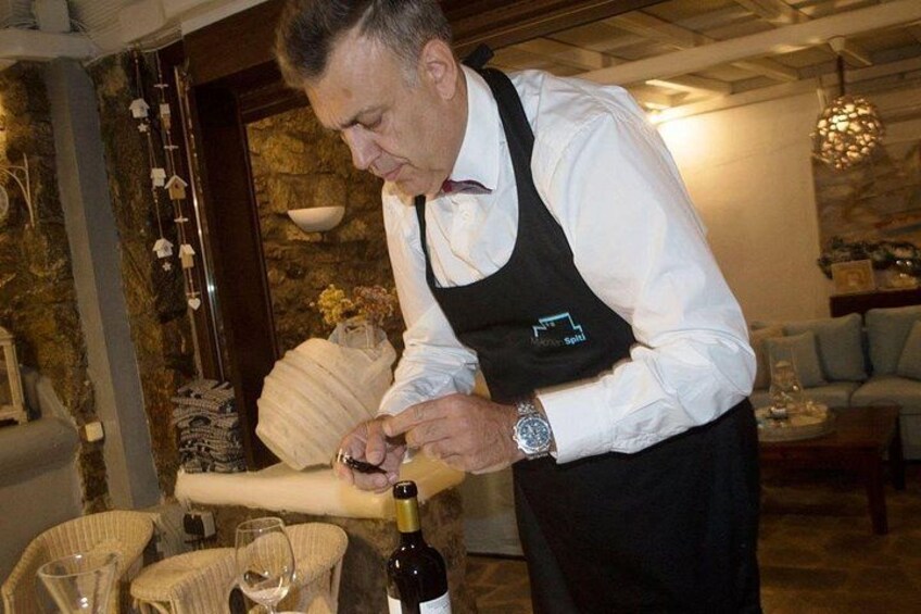 A person in a white shirt and black apron preparing food, with a bottle of wine and a basket of bread on the table. 2