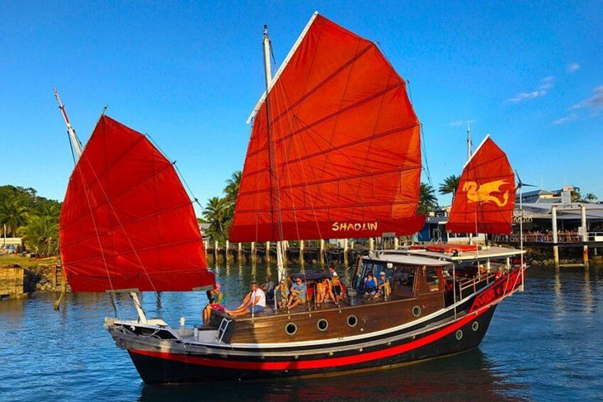 A traditional boat with red sails named 'SHAOLIN' on a calm water body, surrounded by a clear sky and palm trees. 2