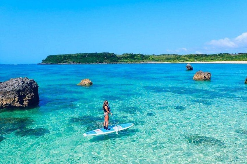 Une personne fait de la planche à pagaie sur une eau turquoise et limpide, avec des rochers et une étendue de terre verdoyante en arrière-plan. 2