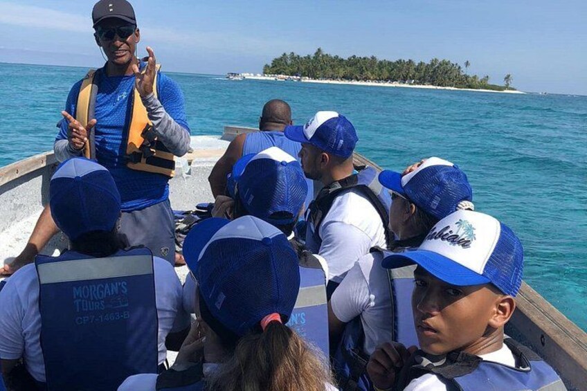 Un groupe de personnes se trouve sur un bateau, portant des gilets de sauvetage et des chapeaux, avec une île tropicale en arrière-plan. 3