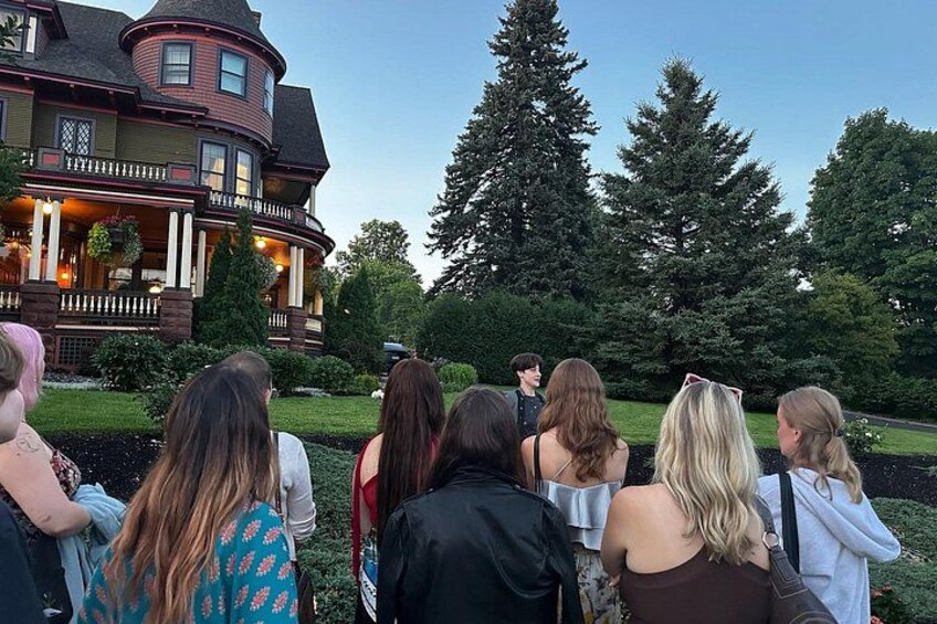 A group of people standing in front of a large, historic house with a porch and a well-maintained garden. 4