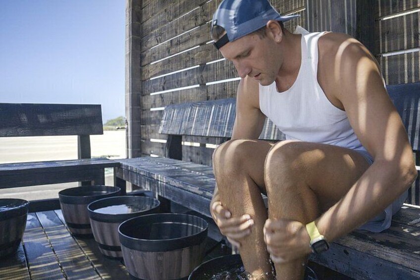 A person sitting on a wooden bench outdoors, with metal buckets and a wooden wall in the background. 3