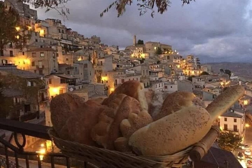 Paesaggio urbano al crepuscolo con edifici illuminati da lampioni, un cesto di pane in primo piano e una catena montuosa sullo sfondo. 4
