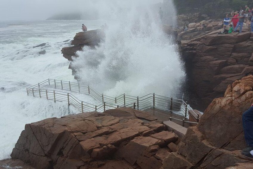 Eine felsige Küste mit einer Treppe, die zum Meer führt, Wellen schlagen an den Felsen und Menschen beobachten die Szene. 2