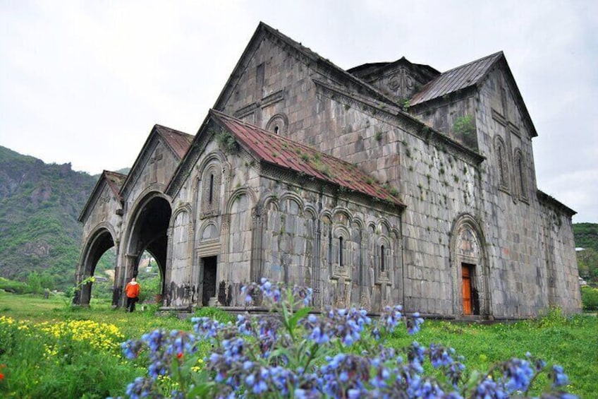 Eine alte Steinkirche mit Bögen und einem Glockenturm, umgeben von grünem Gras und Wildblumen. 1