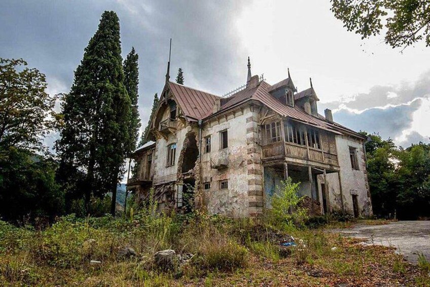 An abandoned, dilapidated house with a steep roof and stone walls, surrounded by overgrown vegetation and tall trees. 2