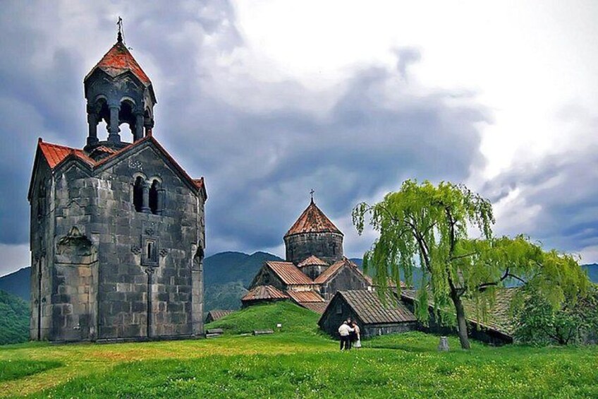 A stone church with a bell tower and a smaller building with a red roof, surrounded by lush greenery and a cloudy sky. 5
