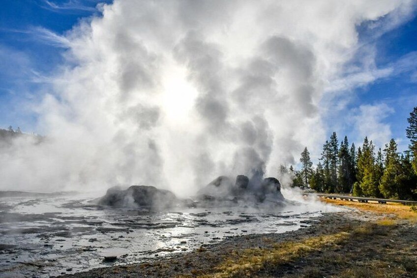 A geyser eruption with steam, a forested area, and a paved path. 2
