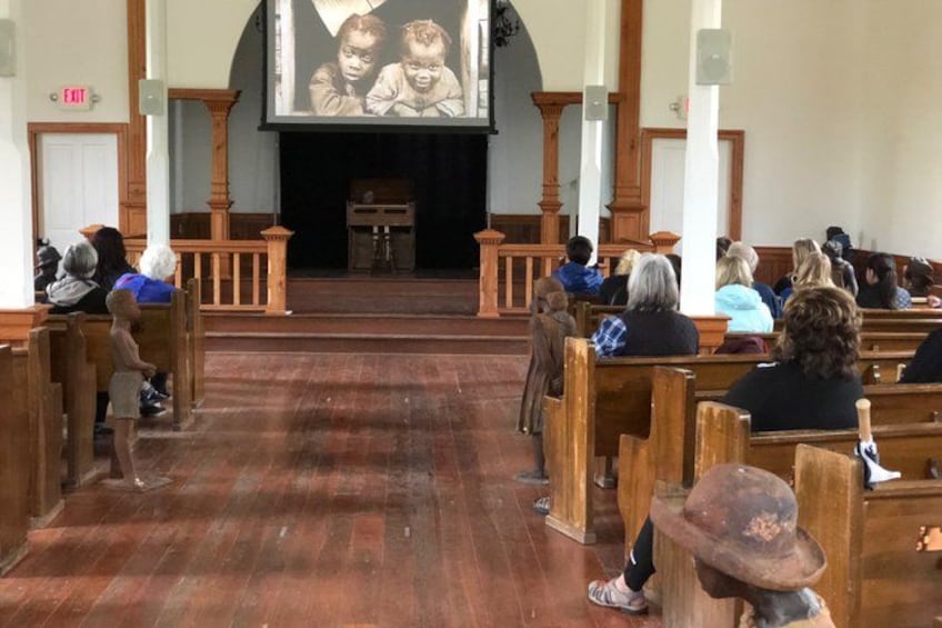 Ein Kircheninneres mit Holzbänken, einem hölzernen Altar und einem Bildschirm, der ein Bild zeigt. 2
