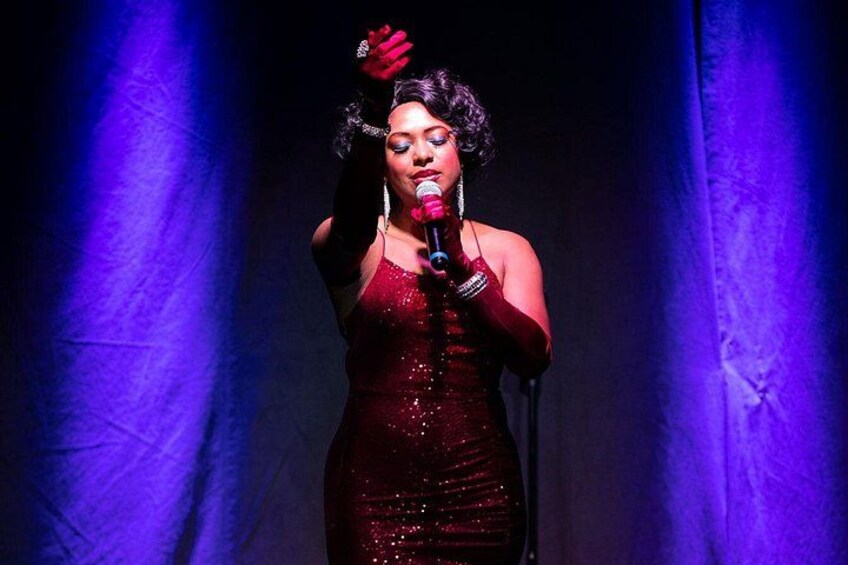 A person in a sparkling red dress holding a microphone on a stage with purple lighting. 1