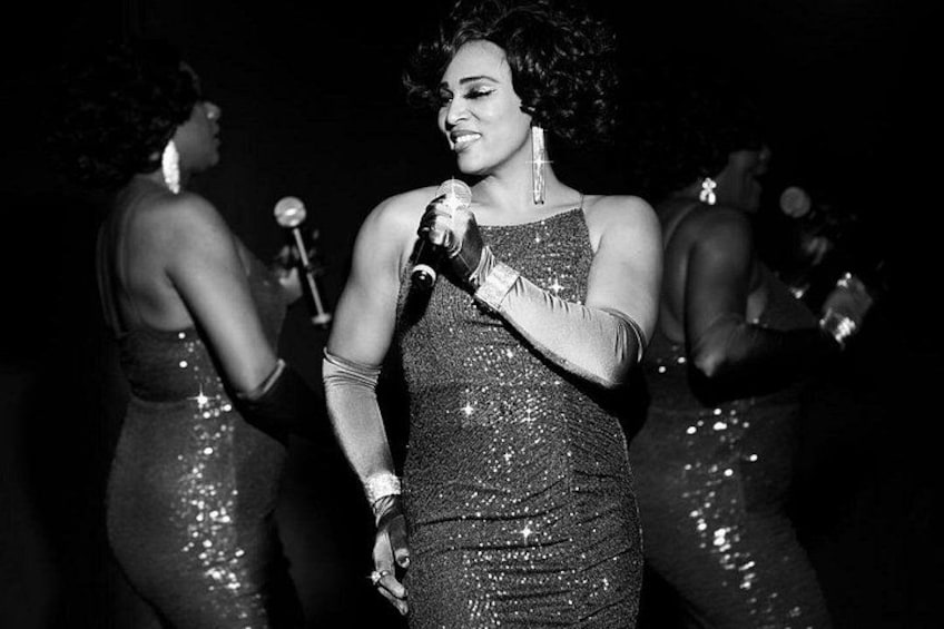 A person in a sparkly dress holding a microphone, with other people in the background. 2