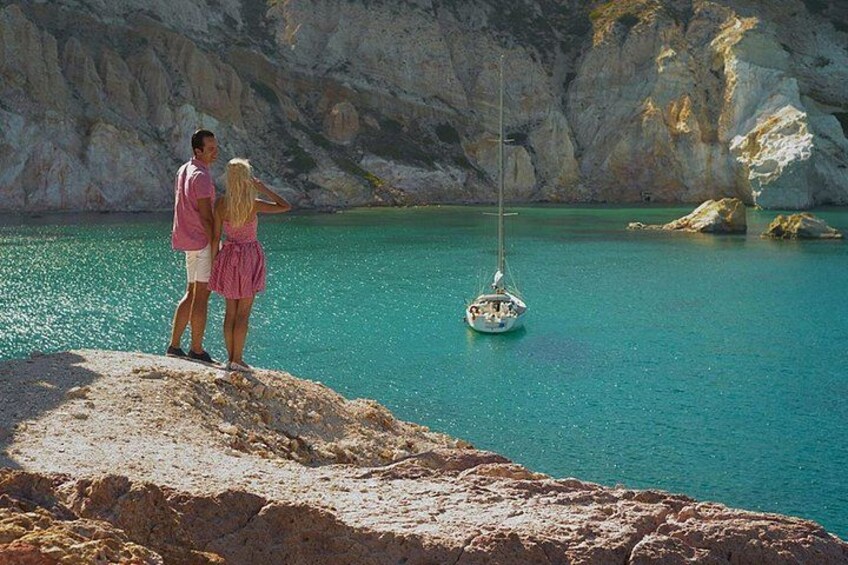 A couple standing on a rocky outcrop overlooking turquoise water with a sailboat in the distance. 4