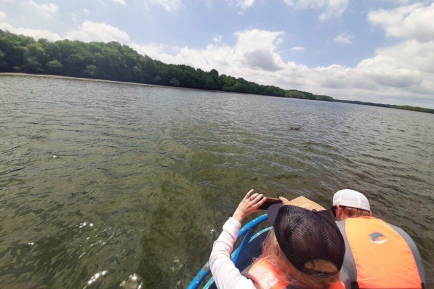 A person in a boat wearing an orange life jacket, another person in a white cap, and a forested shoreline in the background. 4