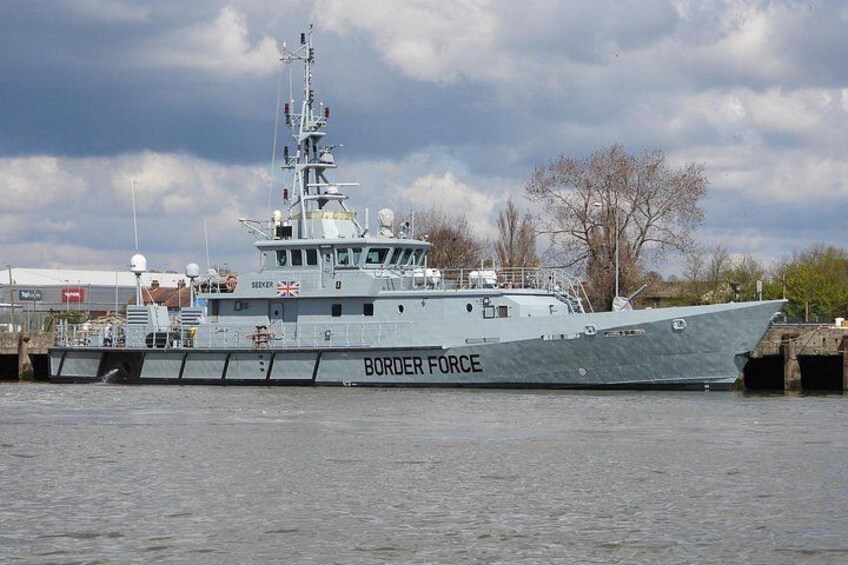 A large naval ship named SEVEN with the word BORDER FORCE on its side, docked at a port with a cloudy sky above. 2