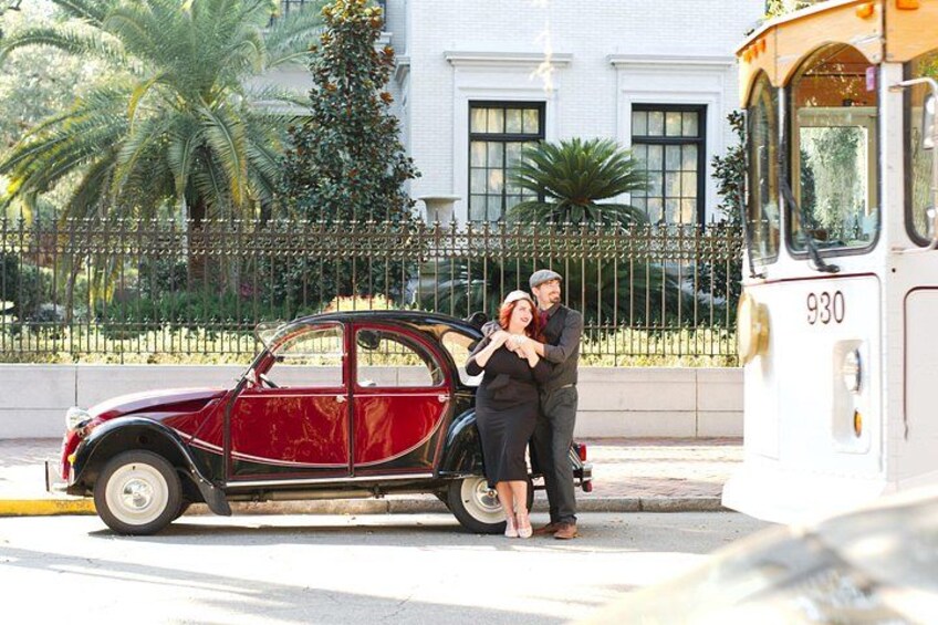 A vintage car parked on the side of a street, a man and a woman embracing, and a bus in the background. 2
