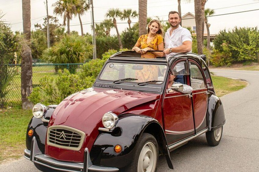 A vintage car parked on a paved road with greenery and palm trees in the background. 3