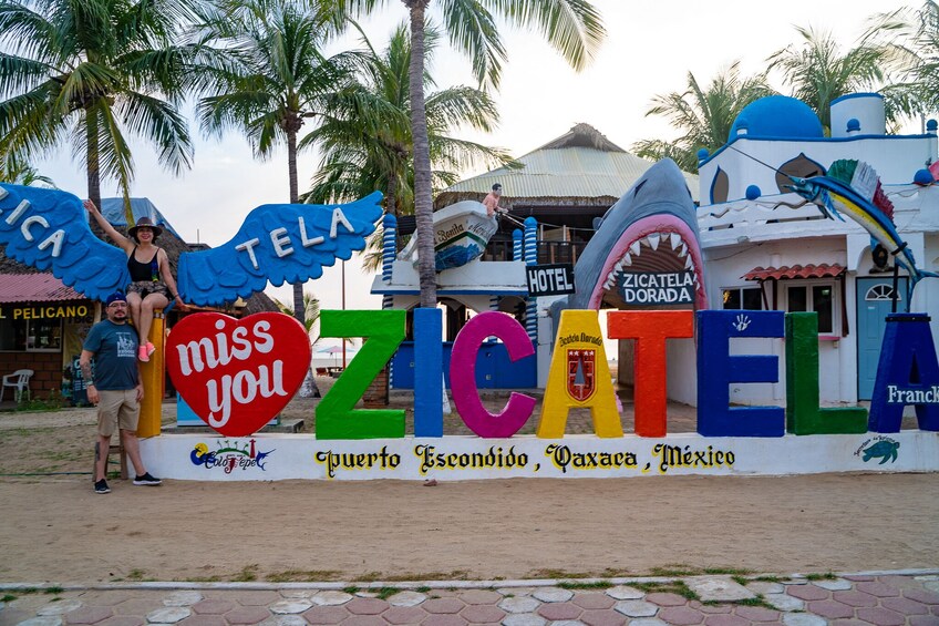 Un letrero colorido con 'ZICATEL' y 'te extraño' en letras grandes, con palmeras y un edificio al fondo. 1
