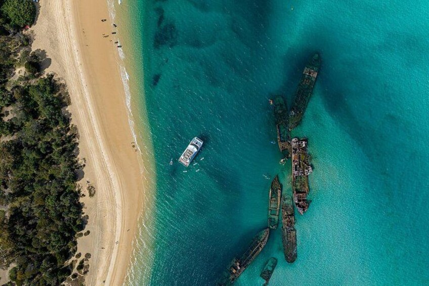 An aerial view of a beach with clear turquoise waters, a small boat, and several sunken ships. 2