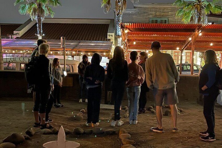 Un grupo de personas reunidas alrededor de un objeto pequeño e iluminado en el suelo, posiblemente una fuente o una escultura, en un entorno exterior con palmeras y un edificio al fondo. 3