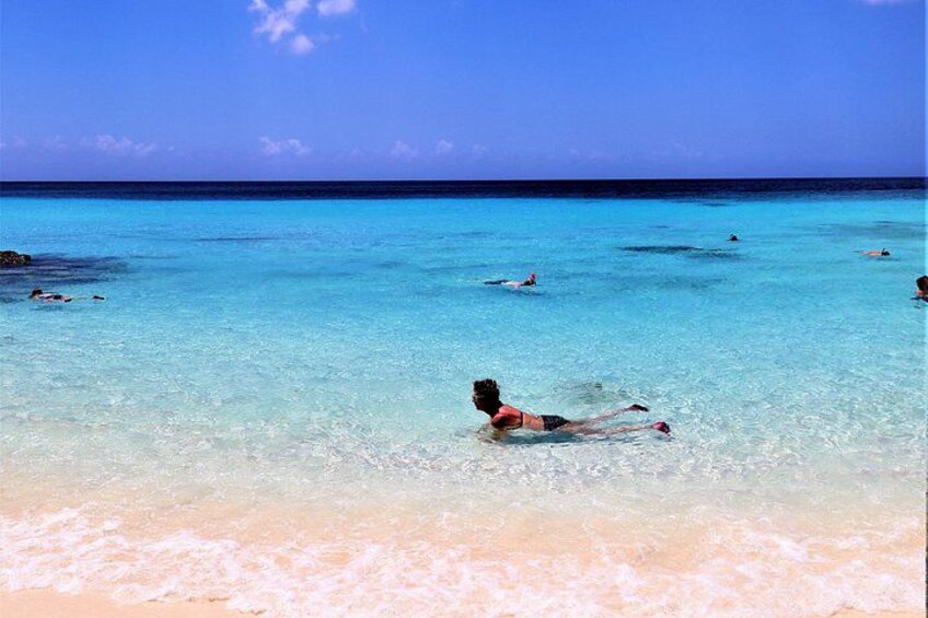 A beach with clear turquoise water, a person lying on the sand, and others swimming in the distance. 2
