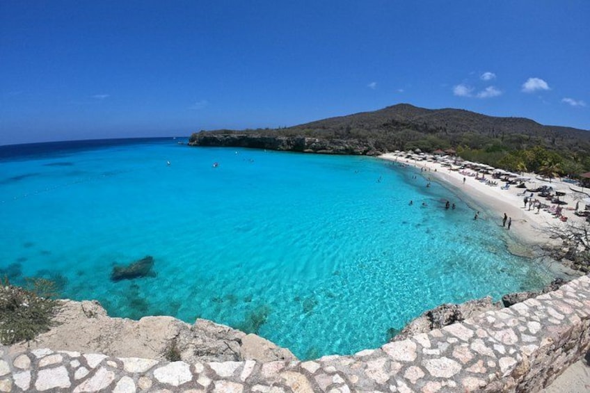 Ein türkisfarbener Strand mit klarem Wasser, einer Steinmauer und einem Bergpanorama im Hintergrund. 1