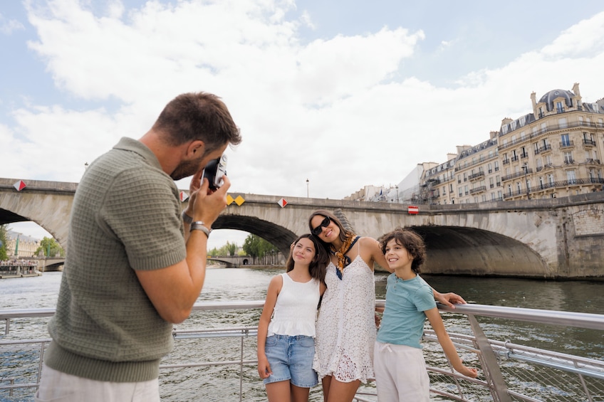 A group of people posing for a photo in front of a historic bridge with a river and a building in the background. 1