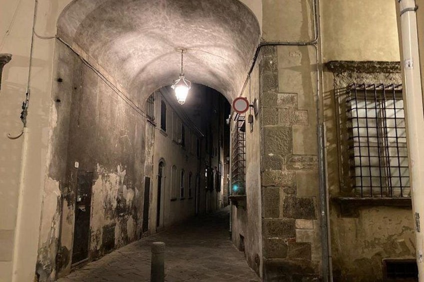 A dimly lit, narrow alleyway with a curved ceiling, stone walls, and a barred window. 5