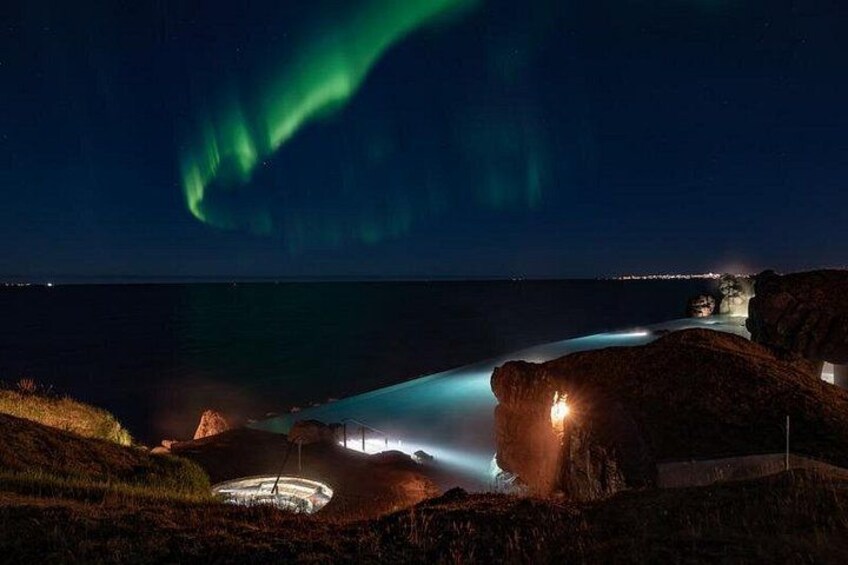 Paesaggio costiero notturno con un'aurora verde, una fontana illuminata e una linea di costa rocciosa. 5