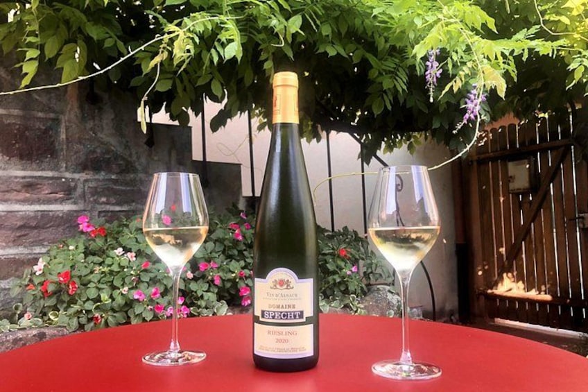 A bottle of Riesling wine and two glasses on a red table, surrounded by greenery and flowers. 3