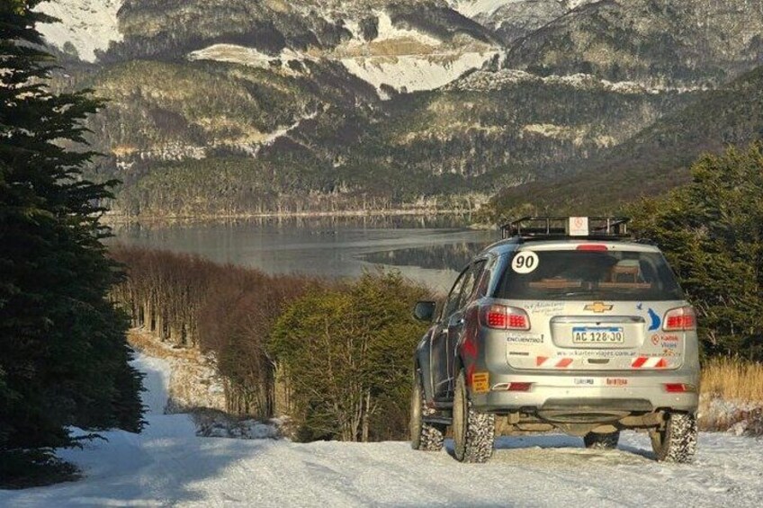 Un vehículo con el número 90 en la parte trasera, estacionado en un sendero cubierto de nieve, con un lago y un bosque al fondo. 2