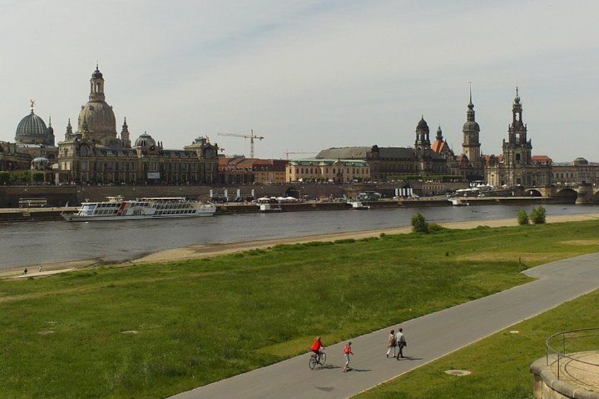 A river with boats, a grassy area, a paved path, and historic buildings in the background. 2