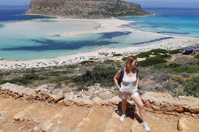 A person standing on a rocky outcrop overlooking a coastal landscape with a beach, clear blue waters, and a distant island. 2