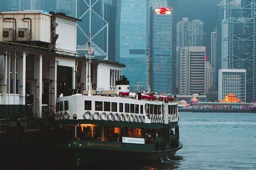 A ferry with a white upper deck and green lower deck, docked at a pier with a city skyline in the background. 1