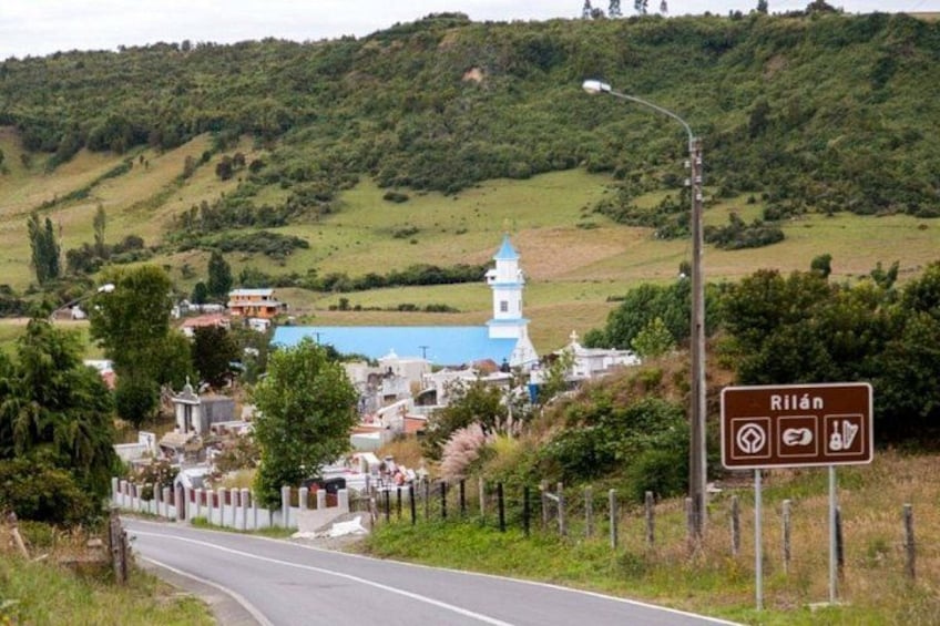 Une route menant à une ville où se trouvent une église et un panneau indiquant « Rilán  », avec des symboles représentant une église et des toilettes. 1