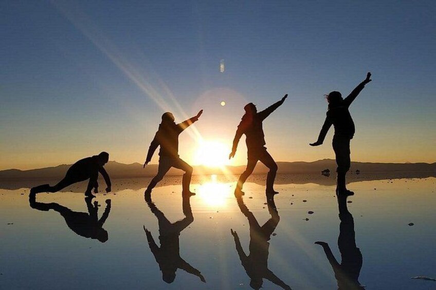 Des silhouettes de personnes dansant sur une surface réfléchissante, avec le soleil couchant en arrière-plan. 1