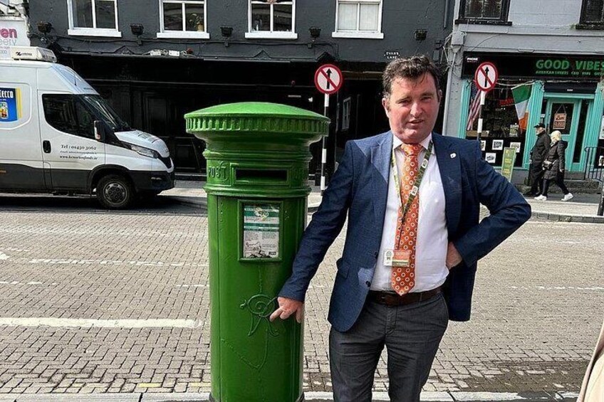 Un hombre con traje azul de pie junto a un buzón verde en la calle. 1