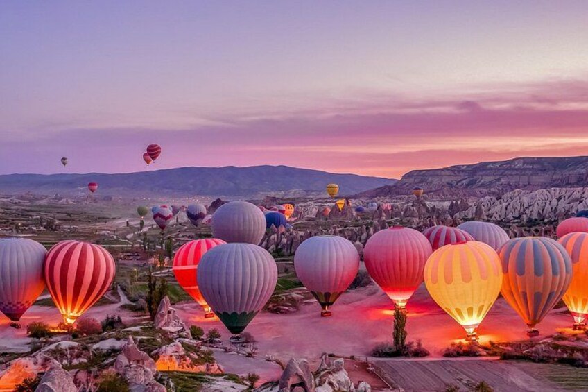 Eine Landschaft mit zahlreichen Heißluftballons, einige davon beleuchtet, und einer bergigen Kulisse. 3