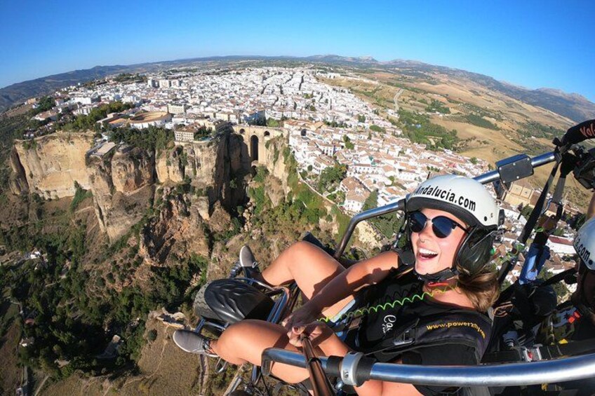 A person in a parachute harness with a helmet labeled 'Andalucia.com' and a website 'www.powerx.com' on the back, flying over a town with a cliffside and a bridge. 1