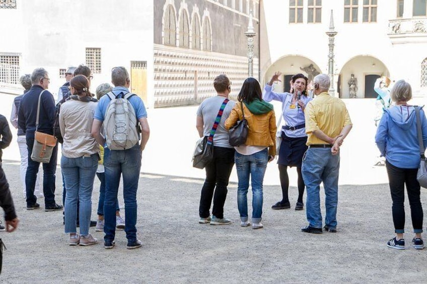 Un groupe de touristes se tient dans une cour, devant une grande fresque murale, avec une architecture classique en arrière-plan. 1