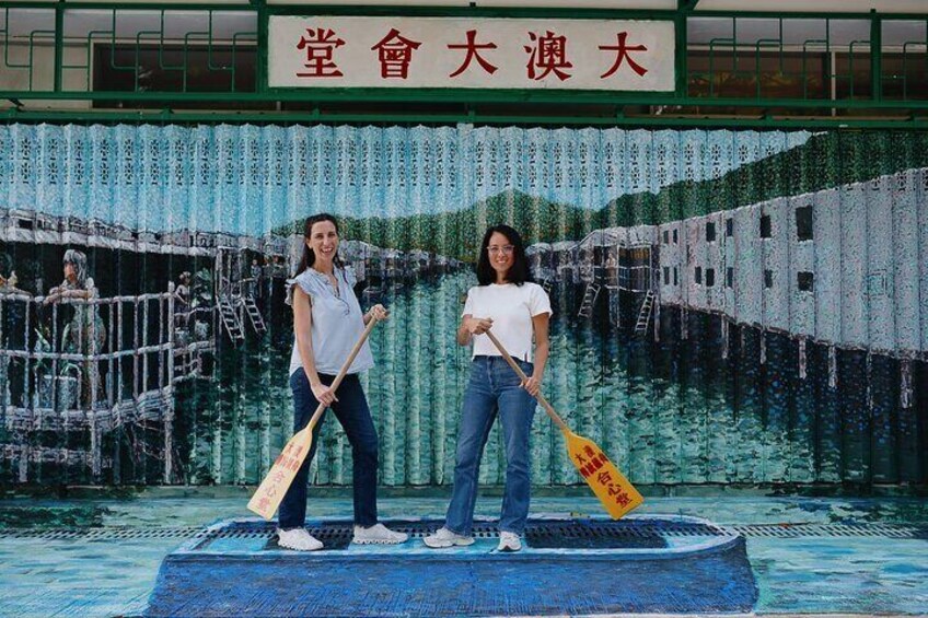 Two people standing on a stage with a backdrop of a painted scene, holding paddles. 3