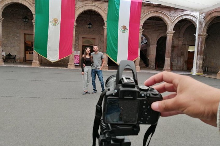 A couple standing in front of a building with arches and two flags. 2