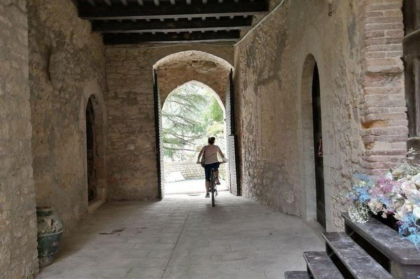 Una persona pedalea en bicicleta por un corredor de piedra con arco, con un banco a la derecha. 1