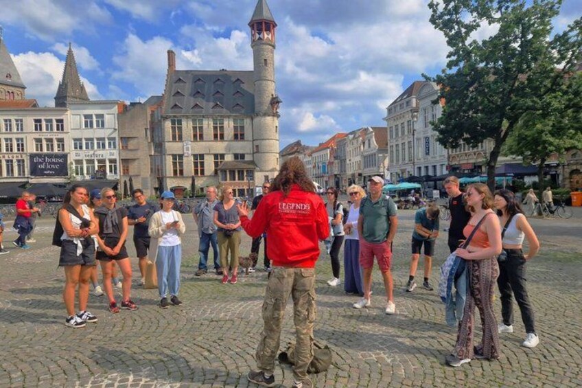 A group of people standing in a cobblestone square with historic buildings and a tower in the background. 1