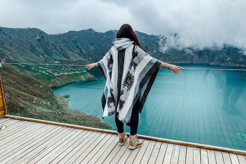 Eine Person steht auf einem Holzboden, die Arme ausgebreitet, und blickt auf einen türkisblauen See und eine gebirgige Landschaft. 1