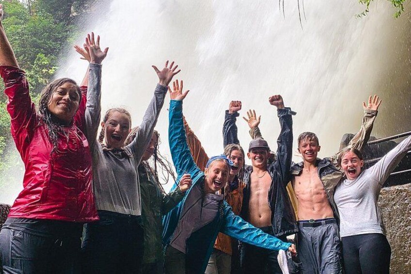 Eine Gruppe von Menschen steht vor einem Wasserfall, die Arme erhoben. 4