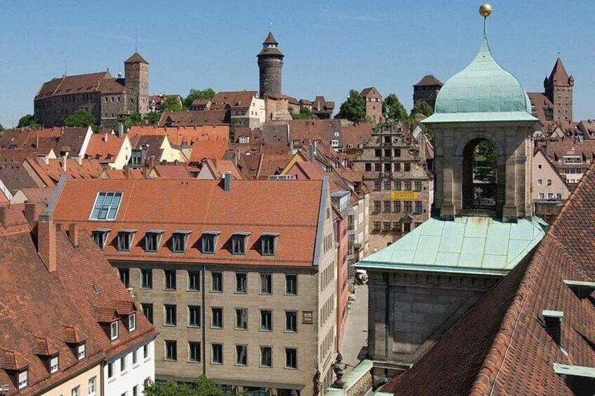Une ville historique, avec des bâtiments aux toits rouges, une tour imposante et une église surmontée d’un dôme vert. 1